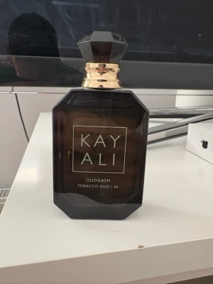 Kayali Oudgasm Tobacco Oud parfym - Kayali Oudgasm Tobacco Oud 04 är en lyxig parfym i en mörkbrun, nästan svart, fyrkantig glasflaska med gulddetaljer och en elegant svart facettslipad kork. Doften har tydliga inslag av oud och tobak, vilket ger en djup och mystisk känsla.