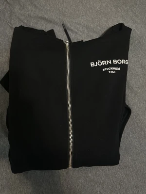 Svart zip hoodie från Björn Borg - Säljer en svart hoodie från Björn Borg med dragkedja framtill och vit logga på bröstet. Hoodien har huva med dragsko och långa ärmar. Perfekt för en avslappnad och sportig stil.