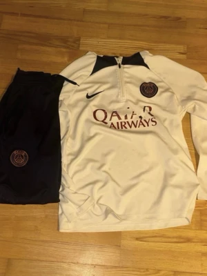 PSG träningsset Nike vit/svart - Säljer ett snyggt och skönt träningsställ från Paris Saint-Germain (PSG) från Nike. Setet innehåller både den långärmade träningströjan (Dri-FIT Strike Drill Top) och de matchande träningsbyxorna.  Märke: Nike / Paris Saint-Germain Storlek: S men Passar M oxå både byxor och tröjan  Skick: Bra begagnat skick. Tyget och passformen är i jättefint skick, men "Qatar Airways"-loggan på bröstet har släppt lite i tvätten (se sista bilden för detaljer).Perfekt till fotbollsträningen eller som sköna mjuki