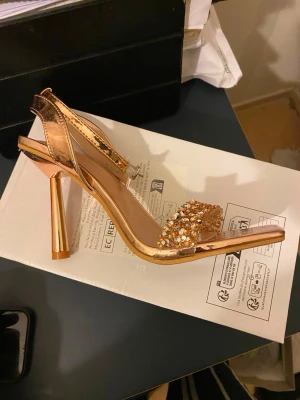 Glittriga guldiga pumps med rem - Säljer ett par glittriga pumps i guld med fyrkantig tå och transparent rem över foten. Klacken är hög och i metallic guld, och framtill finns dekorativa paljetter och stenar. Snygg rem runt ankeln med spänne. Perfekta för dig som vill sticka ut.