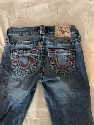 True Religion blå jeans med röd söm - Säljer ett par blå jeans från True Religion med ikoniska röda kontrastsömmar och detaljerade bakfickor. Klassisk femficksmodell med silverfärgade knappar och patch bak i midjan. Perfekta för dig som gillar streetwear och vill sticka ut.