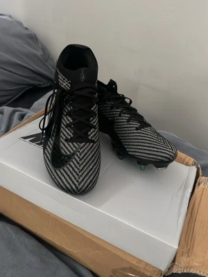 Svarta och gråa Nike fotbollsskor - Säljer ett par snygga fotbollsskor från Nike i svart och grått med ett mönstrat stickat material. Skorna har snörning, hög krage för extra stöd och klassisk Nike-logga på sidan. Perfekta för dig som vill ha stilrena och funktionella skor på planen.