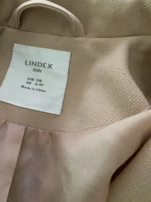 Beige kappa från Lindex Kids - Stilren beige kappa från Lindex Kids i storlek 134. Kappan har klassisk krage och är helfärgad med mjukt foder på insidan. Perfekt för dig som vill ha en enkel och snygg look till hösten eller våren.