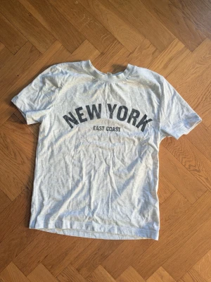 Grå t-shirt med New York-tryck - Snygg grå t-shirt med svart text 'NEW YORK EAST COAST' på bröstet. Klassisk rund hals och korta ärmar. Perfekt för dig som gillar streetstyle och vill ha en enkel men trendig look.
