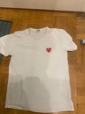 Cdg T-shirt  - Vit t-shirt med rund hals och korta ärmar. På bröstet sitter ett litet rött hjärta med ögon, vilket ger en unik och lekfull touch. Perfekt för dig som gillar enkel men ändå personlig stil.