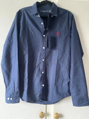 Marinblå skjorta från Ralph Lauren - Klassisk marinblå skjorta från Ralph Lauren med röd broderad logga på bröstet. Skjortan har långa ärmar, knappar framtill och är tillverkad i mjuk bomull. Perfekt för en stilren och avslappnad look.