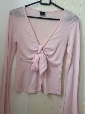 Ljusrosa långärmad topp med knyt - Söt ljusrosa långärmad topp med djup v-ringning och knyt framtill. Tillverkad i ett tunt, mjukt material som ger en feminin och trendig look. Perfekt att styla med jeans eller kjol för en avslappnad men snygg outfit.