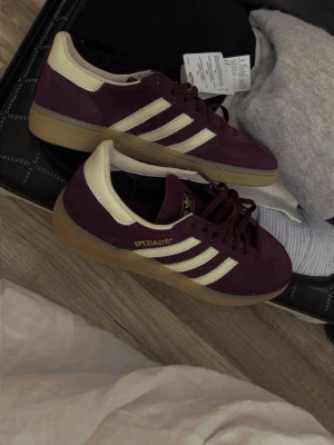 Adidas Spezial vinröda sneakers - Säljer ett par Adidas Spezial sneakers i vinrött mocka med benvita detaljer på sidorna och beige hälparti och gummisula. Snygg retrostil som passar perfekt till streetwear och casual outfits. Prislappen är tyvärr borttagen men dem är i nyskick. Fler bilder finns om intresse finns💕 Kommer levereras i kartong så inget händer. Säljer för att dem inte kommer till användning, gör lite ont k hjärtat men det är bättre att de hamnas hos dig som använder de☹️💕 (ord pris 1200kr)