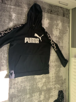 Svart Puma hoodie med logga - Svart hoodie från Puma med stor vit logga och text på bröstet. Huva med dragsko och magficka framtill. Ärmarna har vita Puma-detaljer längs hela sidan för en sportig vibe. Perfekt för chill eller streetwear.