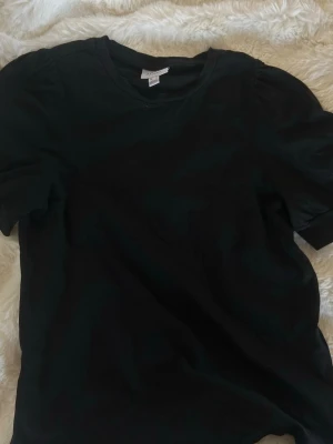 Svart puffärmad t-shirt - Säljer en svart t-shirt med puffärmar och rund hals. T-shirten har en enkel och clean look, perfekt till jeans eller kjol. Materialet känns mjukt och skönt, troligtvis bomull. Passformen är normal och modellen är klassisk.