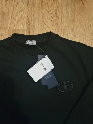 Svart sweatshirt från Dior - Svart sweatshirt från Dior med rund halsringning och diskret broderad Dior-logga på bröstet. Tröjan har långa ärmar och ribbade muddar vid ärmslut och nederkant. Perfekt för en stilren och avslappnad look.