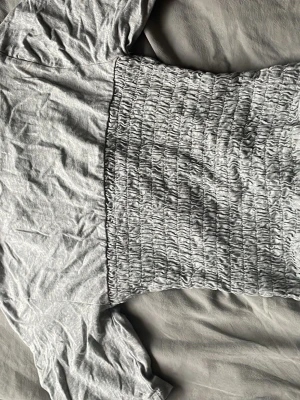 Grå smockad topp från H&M Divided - Säljer en ljusgrå smockad topp från H&M Divided. Toppen har korta puffärmar och en tight, smockad midja som ger en snygg siluett. Tillverkad i mjuk bomull, perfekt för dig som gillar en enkel men trendig stil.