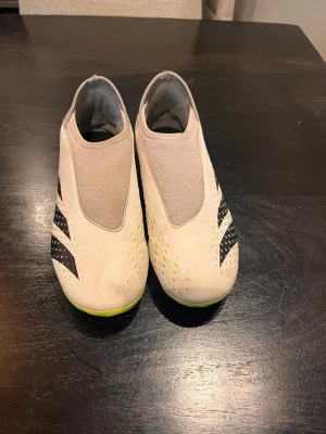Adidas Predator vita fotbollsskor - Säljer ett par vita Adidas Predator fotbollsskor med svarta detaljer och gula inslag. Skorna har en strömlinjeformad design utan snörning och en grön/gul sula för bra grepp på planen. Tillverkade i syntetmaterial och perfekta för dig som vill ha kontroll och stil på fotbollsplanen.