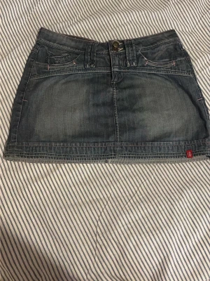 Blå jeanskjol med fickor - Säljer en klassisk blå jeanskjol i kort modell med fram- och bakfickor samt bälteshällor. Kjolen har en knapp och dragkedja framtill och detaljer i kontrastsömmar. Perfekt för dig som gillar en enkel och tidlös denimlook.
