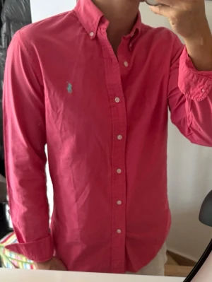 Rosa skjorta med knappar - Säljer en klassisk rosa skjorta med button-down krage och vita knappar. Skjortan har lång ärm och en broderad logga på bröstet. Perfekt för dig som vill ha en stilren och färgglad look. Liten tråddragning på axeln som man ser på sista bilden, därav priset 