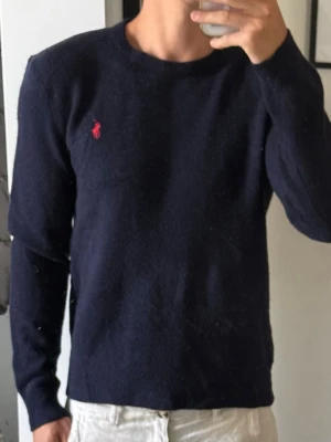 Mörkblå stickad tröja Polo Ralph Lauren - Klassisk mörkblå stickad tröja från Polo Ralph Lauren med rund halsringning och röd broderad logga på bröstet. Tröjan har långa ärmar och ribbade muddar vid ärmslut och nederkant. Perfekt till jeans eller chinos. Storlek m men sitter som S