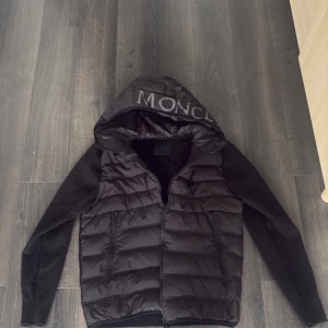 Moncler jacka - Snygg svart pufferjacka med vadderad front och huva där det står 'MONCLER. Jackan har dragkedja framtill, två sidofickor med dragkedja och släta svarta ärmar. Perfekt för kalla vinterdagar och ger en modern look.
