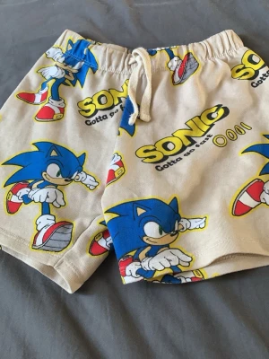 Beiga Sonic shorts med snörning - Säljer ett par beiga shorts med färgglatt Sonic-motiv och texten 'Gotta go fast'. Shortsen har snörning i midjan och elastisk resår för skön passform. Perfekta för dig som gillar gaming och vill ha något bekvämt och lekfullt i garderoben.