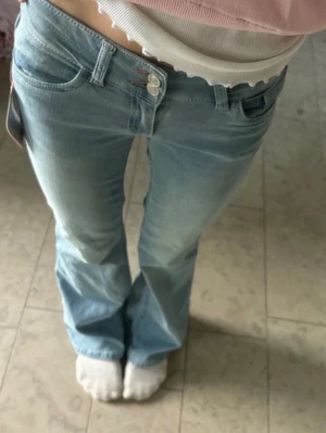 Ljusblå bootcut jeans med rosa detaljer - Säljer ett par ”low waist bootcut slitch jeans”  från Nelly. Fickorna och knapparna har broderat rosa.💕