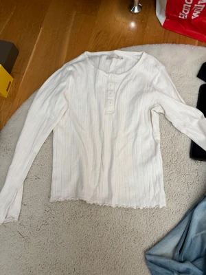 Vitt ribbat set med långärmad topp - Säljer ett vitt ribbat set med långärmad topp och matchande byxor. Toppen har knappar framtill och en lätt vågig kant nertill. Byxorna har resår i midjan och vida ben. Materialet är mjukt och stretchigt, perfekt för att chilla hemma.