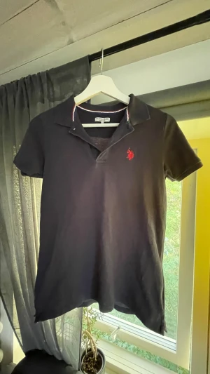 Mörkblå pikétröja från U.S. Polo Assn - Klassisk svart pikétröja från U.S. Polo Assn med korta ärmar och röd logga broderad på bröstet. Kragen har en snygg detalj med röd, vit och blå rand. Perfekt till jeans eller shorts för en clean look. Storlek 12-13 år