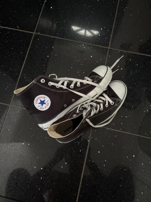 Svarta Converse Chuck Taylor High - Klassiska svarta Converse Chuck Taylor All Star high-tops med vit tåhätta och vita snören. Skorna har den ikoniska loggan på sidan, vit sula och kontrastsömmar. Tillverkade i canvas med gummidetaljer. Perfekta för en avslappnad och cool stil.