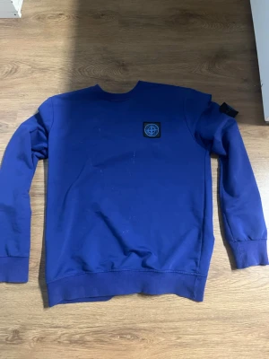Blå sweatshirt från Stone Island - Snygg blå sweatshirt från Stone Island med klassisk patch på ärmen. Tröjan har rund halsringning, ribbade muddar och en avslappnad passform. Perfekt för dig som gillar streetwear och vill ha en stilren look.