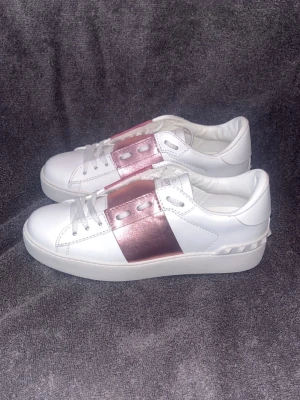 Valentino vita sneakers med rosa band - Snygga vita sneakers från Valentino med ett metallicrosa band över snörningen. Skorna har vita snören, låg profil och coola nitar på hälen. Tillverkade i slätt läder för en clean och modern look.