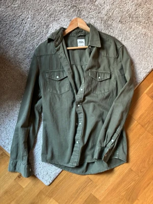 Grön skjorta från Zara med knappar - Säljer en grön skjorta från Zara med tryckknappar och två bröstfickor. Skjortan har klassisk krage och lång ärm, perfekt för en avslappnad men snygg look. Materialet känns mjukt och bekvämt, troligen bomull.