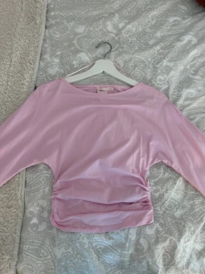 Ljusrosa topp med rynkade detaljer - Säljer en ljusrosa topp med långa ärmar och snygga rynkade detaljer i midjan. Toppen sitter tajt i midjas med utsvängda armar ner till. Perfekt för dig som vill ha mer färg i din vardag! 