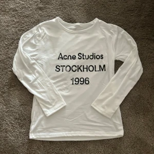 Vit långärmad topp Acne Studios - Vit långärmad topp från Acne Studios med tryck i svart text på bröstet där det står 'Acne Studios STOCKHOLM 1996'. Klassisk rund halsringning och mjukt material som känns skönt mot huden.