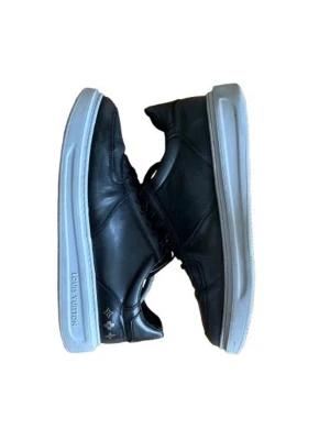 Svarta Louis Vuitton sneakers i läder - Svarta sneakers från Louis Vuitton i slätt läder med vita sulor och diskreta logodetaljer på sidan och plös. Klassisk låg modell med svarta skosnören och perforerad tå. Perfekta för dig som gillar stilrena och exklusiva sneakers.
