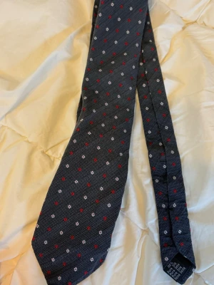 Mörkblå slips med småmönster - Snygg mörkblå slips med små vita och röda blommönster. Slipsen har klassisk form och är tillverkad i ett tyg med diskret struktur. Perfekt för dig som vill addera lite färg och mönster till din outfit.