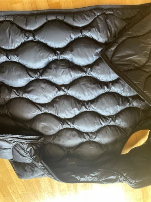 Svart dunväst från Peak Performance - Snygg svart dunväst från Peak Performance med vågformat quiltat mönster och dragkedja framtill. Västen är lätt och har en sportig look, perfekt för lager-på-lager. Tillverkad i glansigt material som ger en modern vibe.