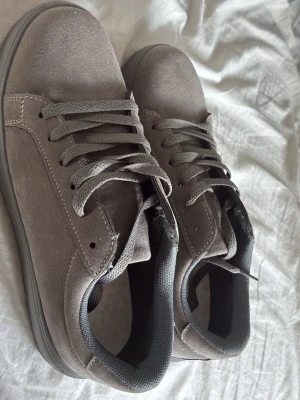 Gråa låga sneakers i textil - Säljer ett par stilrena gråa sneakers i textil med matchande grå skosnören och sula. Skorna har en klassisk låg design och diskreta sömmar som ger en clean look. Perfekta för dig som gillar enkel och snygg stil.