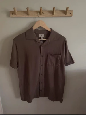 Kortärmad skjorttröja HM - Helt ny med prislapp kvar. Det är en skjorttröja från HM med väck längst ner på sidorna (se den sista bilden). Den är ganska tunn, perfekt för sommar. Färgen är en slags brun/grå nyans.   Passformen är relaxed fit. Jag skulle säga att den sitter som en S.   Nypris 399 kr.   Kontakta gärna för frågor!
