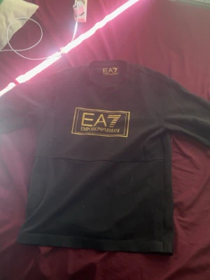 Svart EA7 Emporio Armani sweatshirt - Snygg svart sweatshirt från EA7 Emporio Armani med guldfärgat tryck på bröstet. Tröjan har rund halsringning och långa ärmar. Perfekt för dig som gillar stilrena och exklusiva plagg med tydlig logga.