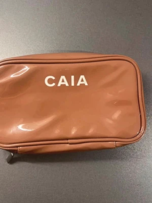 Brun necessär från CAIA - Säljer en brun necessär från CAIA med dragkedja och flera nätfickor inuti. Perfekt för att organisera smink eller småprylar. Stilren design med CAIA-logga på framsidan och praktisk form som är lätt att ta med sig. Mycket sminkfläckar inuti men övrigt mycket fin och hel.