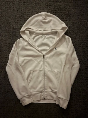 Vit hoodie från Polo Ralph Lauren - Klassisk vit hoodie från Polo Ralph Lauren med dragkedja framtill och huva. Hoodien har två fickor på magen och den ikoniska Polo-loggan broderad på bröstet. Tillverkad i mjuk bomull för en skön och avslappnad stil.