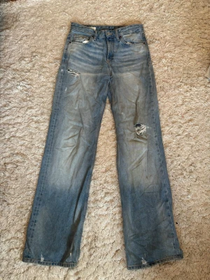 Blå raka jeans med slitningar - Ett par blå jeans med raka ben och tydliga slitningar på låret och knät. Jeansen har klassisk femficksdesign och är gjorda i denim. Perfekta för dig som gillar en avslappnad och trendig stil. Medelhög midja