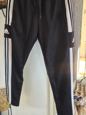 Svarta Adidas träningsbyxor med vita ränder - Säljer ett par svarta träningsbyxor från Adidas med klassiska vita ränder längs sidorna och Adidas-logga på låret. Byxorna har dragsko i midjan och är tillverkade i ett mjukt, stretchigt material som passar perfekt till träning eller chill.