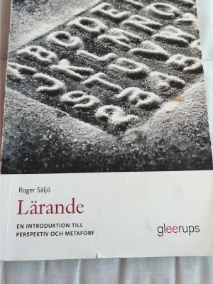 Lärande av Roger Säljö - Kursbok med titeln 'Lärande: En introduktion till perspektiv och metafor' av Roger Säljö. Omslaget är vitt med en bild av bokstäver i sand och text i vinrött och svart. Utgiven av Gleerups, perfekt för dig som pluggar pedagogik eller utbildningsvetenskap.