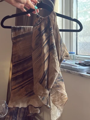 Beige och brun mönstrad sjal - Säljer en tunn och luftig sjal i beige och bruna toner med abstrakt mönster. Sjalen har en rektangulär form och är perfekt att slänga över axlarna för en cool och avslappnad look. Materialet känns lätt och skönt mot huden.