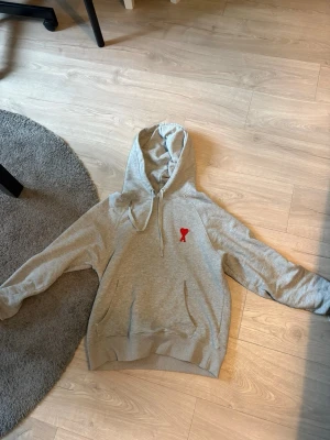Grå ami Paris hoodie - Snygg grå hoodie med huva och dragsko. På bröstet finns en liten röd broderad detalj som sticker ut. Hoodien har känguruficka framtill och ribbade muddar vid ärmslut och nederkant. Perfekt för en chill och avslappnad stil. Storlek M men skulle säga den passar S bättre