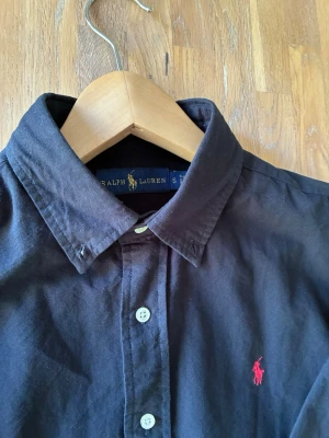 Ralph lauren skjorta mörkblå - Klassisk mörkblå skjorta från Ralph Lauren med knappar framtill och röd broderad logga på bröstet. Skjortan har lång ärm, button down-krage och är tillverkad i mjuk bomull. Perfekt för en stilren och avslappnad look. Skjortan är välldigt mörkblå och kan nästan se ut att vara svart som på bild 2.