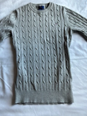 Ljusgrå kabelstickad tröja - Ljusgrå kabelstickad tröja med rund halsringning och ribbade muddar vid ärmslut och nederkant. Tröjan har ett diskret broderat märke på bröstet och är tillverkad i ett mjukt material som passar perfekt till kyligare dagar.