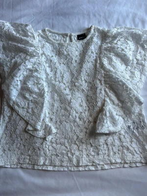 Vit spetsblus med volangärmar - Snygg vit blus i spets med blommigt mönster och stora volanger längs ärmarna. Blusen har rund halsringning och knäppning bak i nacken. Perfekt för dig som gillar romantisk och trendig stil.