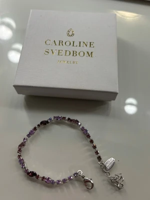 Lila Caroline Svedbom armband - Snyggt armband från Caroline Svedbom med gnistrande pärlor i lila nyanser. Armbandet har en tunn silverfärgad kedja och justerbart lås med liten loggbricka. Helt nytt aldrig använt, säljer då jag inte använder silver smycken längre.