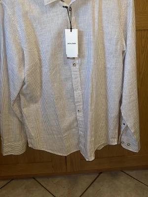 Randig skjorta från Jack & Jones - Säljer en stilren skjorta från Jack & Jones i vitt och ljusbrunt med tunna ränder. Helt ny med prislapp kvar nypris 500 kr