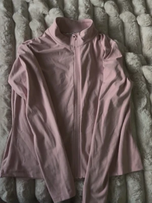 Ljusrosa ziptröja med hög krage - Säljer en ljusrosa ziptröja med hög krage och långa ärmar. Tröjan har en hel dragkedja framtill och är gjord i ett mjukt, stretchigt material. Perfekt för en avslappnad och sportig stil.